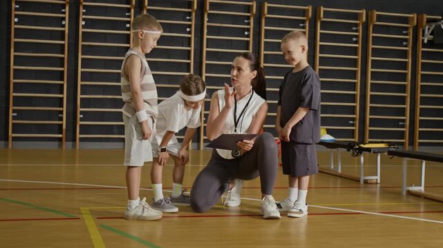 Caucasian female coach kneeling in gym and talking to group of Caucasian kids while holding clipboard. Clip is ideal for school sports programs and physical education marketing
