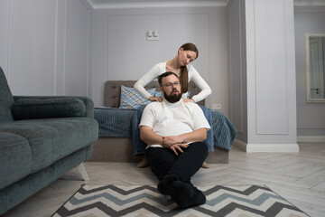 Fototapeta premium Woman standing behind a man sitting on the floor, giving him a shoulder massage to help relieve stress and promote wellness in their cozy home environment