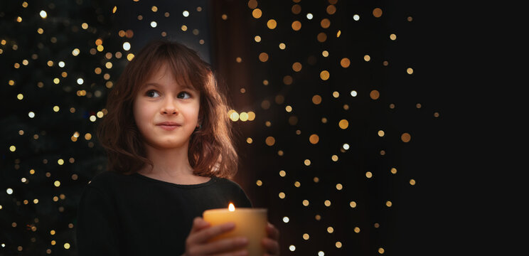  blurred bokeh background and 6-Year-Old Girl Blowing Out Candle Under Christmas Tree in evening near gurlands and wish moment. New year celebrating banner copy space