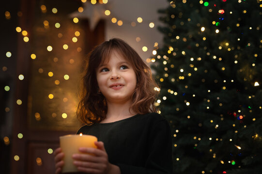 6-Year-Old Girl Blowing Out Candle Under Christmas Tree in evening near gurlands and wish moment. New year celebrating