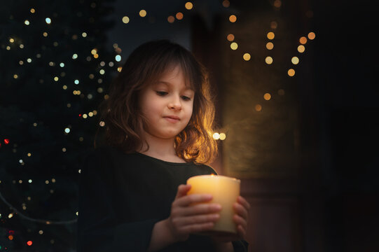 6-Year-Old Girl Blowing Out Candle Under Christmas Tree in evening near gurlands and wish moment. New year celebrating