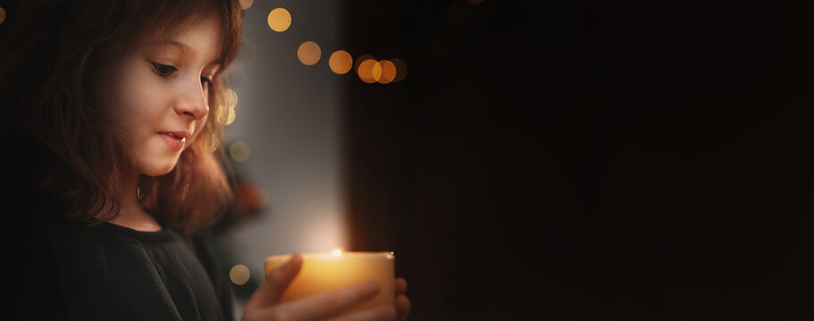 Portrait of 6-Year-Old Girl Blowing Out Candle Under Christmas Tree in evening near gurlands and wish moment. New year celebrating banner copy space