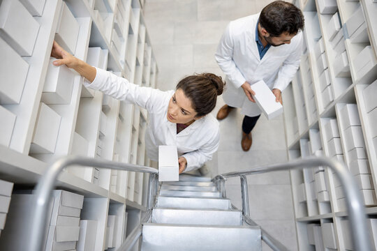 Two scientists in white coats reach for items on high storage shelves, ascending a metal staircase. Concept of inventory, teamwork, and organization in a busy lab or warehouse.