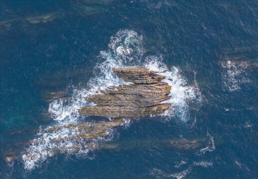 Aerial view of jagged rock formation surrounded by crashing waves and deep blue ocean water in Portugal.