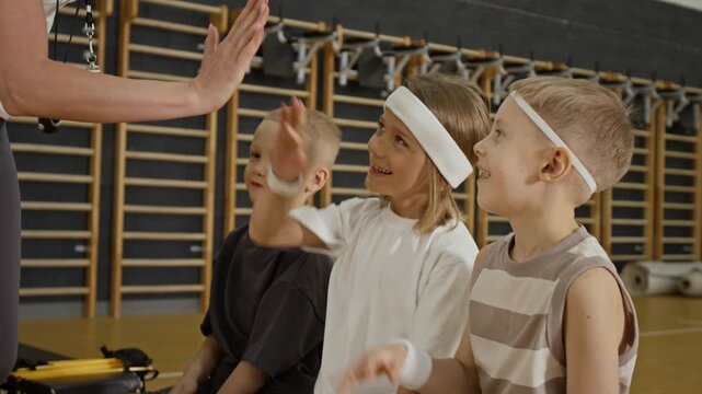 Caucasian children sitting on bench in gym giving high fives to female coach. Perfect for physical education, youth sports, and school fitness commercials