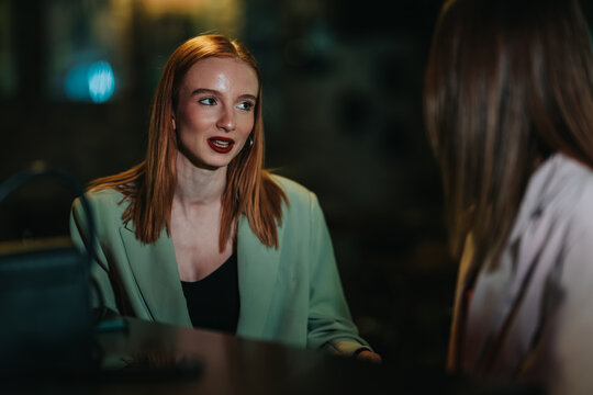 Two young women sit and talk at a dimly lit bar, one redhead speaking to her friend. The scene shows a relaxed evening conversation and social outing between friends.