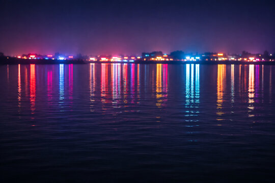 Calm water surface reflecting colorful neon lights from a distant shore.