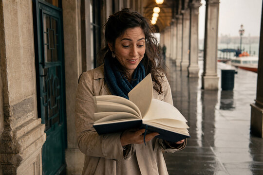 A woman in a beige coat and scarf reads a book along a rainy outdoor colonnade, eyes focused, a gentle smile as pages turn, candid lifestyle moment under cloudy skies.
