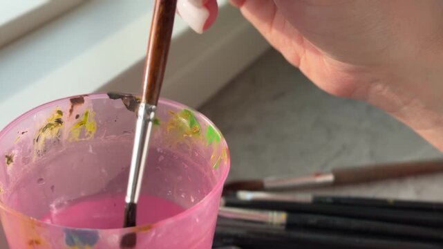 A hand holding a paintbrush above a pink cup with water.