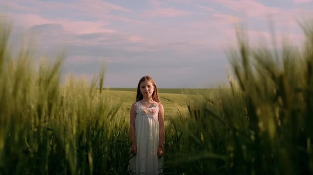 Centered young girl stands calmly in tall green wheat field during golden hour, slight head tilt and relaxed arms conveying peaceful, contemplative mood, with girl, child, wheat, field detail