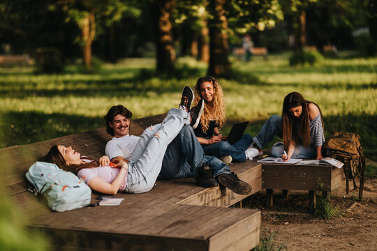 A group of students studying and relaxing on wooden benches in a sunny park. They chat, work on laptops and notebooks while enjoying the outdoor setting.