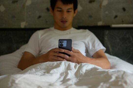 Young Man Using Smartphone in Bed Relaxing with White Bedding