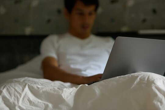 Young Man Working on Laptop in Bed with Cozy White Bedding