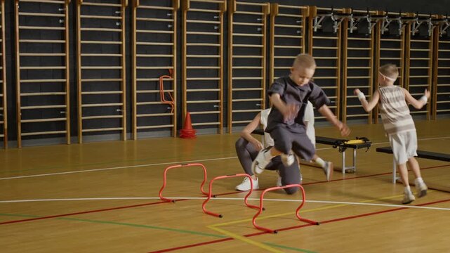 Caucasian kids jumping over hurdles in gym while female Caucasian coach monitoring progress with clipboard. Ideal for physical education promotions, youth sports marketing, and fitness campaigns