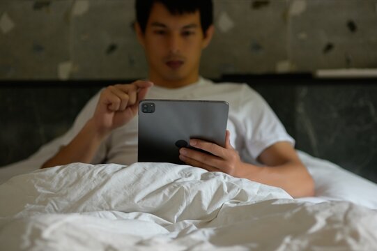 Young Man Relaxing in Bed Using Tablet Device at Home