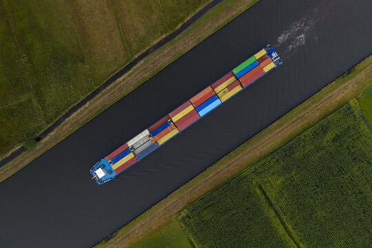 Aerial view of a container barge navigating a narrow canal through green agricultural fields in the Netherlands.