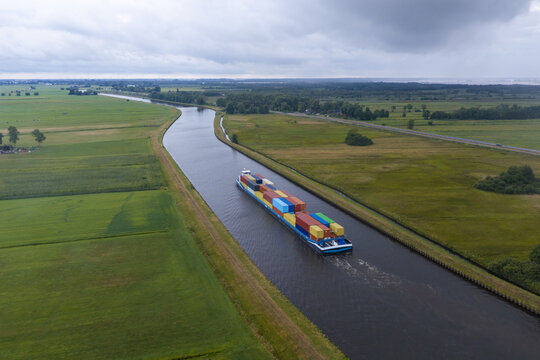 Aerial view of a cargo ship carrying colorful shipping containers navigating through a narrow canal in the rural landscape of the Netherlands.