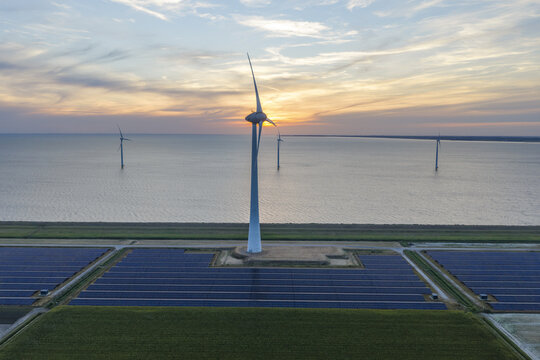 Aerial view of wind turbines standing in the water and rows of solar panels on the shore during a golden sunset in the Netherlands.