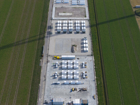 Aerial view of a battery energy storage system facility with rows of white storage containers on a concrete pad surrounded by green agricultural fields.
