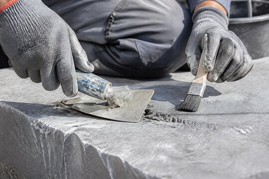 A person wearing gloves is applying a substance to a concrete surface using a trowel and a brush.