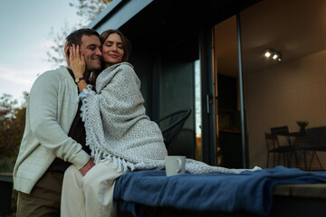 Fototapeta premium Loving couple enjoying a peaceful outdoor moment together, embracing on the patio of their modern home while wrapped in a warm blanket, finding comfort and connection