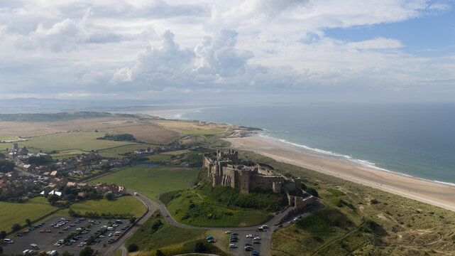Aerial view of Bamburgh Castle situated on a rocky outcrop above the sandy coastline and the North Sea, with green fields and parking areas in Bamburgh, England, United Kingdom.