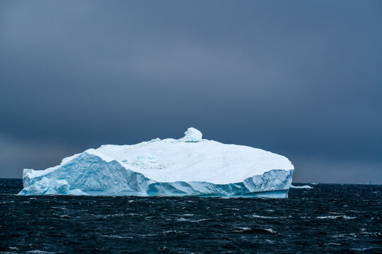 Antarctica: a large white-blue  iceberg floating on a dark sea under an overcast sky.