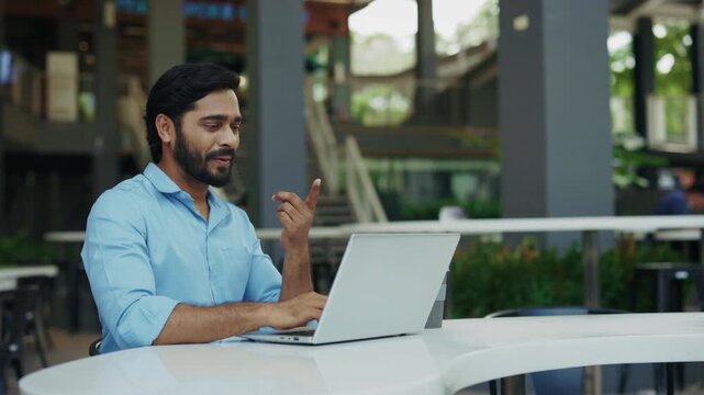 Portrait of friendly Indian entrepreneur talks on video call with sign language in cafe. Interpreter man asks to message back by gestures on laptop during meeting in urban district