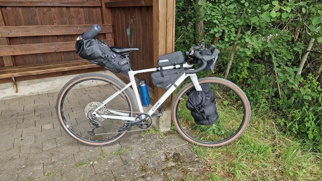 White Cube gravel bike fully loaded with bikepacking bags leaning against a wooden shelter; side view of the gear before a cycling trip in a green setting, Germany.