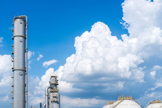 Tall distillation columns and a spherical gas storage tank at a petrochemical plant set against a vivid blue sky with dramatic white cumulus clouds creating a striking contrast.