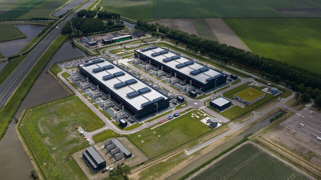 Aerial view of a large data center facility with industrial buildings and flat roofs surrounded by green fields and canals in Middenmeer, North Holland, Netherlands.