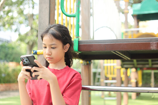 Selective focus outdoor shot captures Asian kid in a red shirt standing at a playground. The young girl is deeply engaged with a smartphone, highlighting modern childhood technology and screen time.