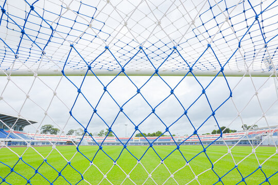 Selective focus of white soccer goal net in the foreground. Through the woven mesh, a blurred empty outdoor football stadium for vibrant green grass pitch under a naturally cloudy daytime sky scene.