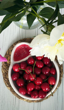 Bowl of fresh red cherries with separate pit section. Summer fruit composition on woven mat with white flower arrangement. Natural rustic food photography on wooden surface