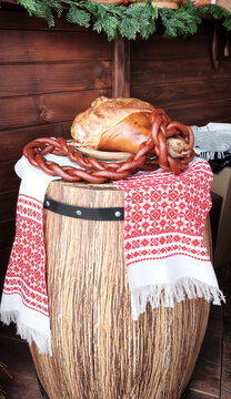 Smoked ham and braided sausage chain on barrel covered with Belarusian embroidered cloth. Rustic festive composition with wood and greenery, symbol of tradition, cuisine, and cultural heritage