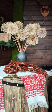 Rustic table with Belarusian folk patterned cloth, chain of sausages arranged decoratively, and vase of dried flowers. Traditional cultural interior with natural textures and cozy atmosphere