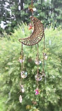 Video of crescent sun catcher with crystals hanging indoors. Rain falls on garden plants in background, creating cozy, magical atmosphere with reflections and nature