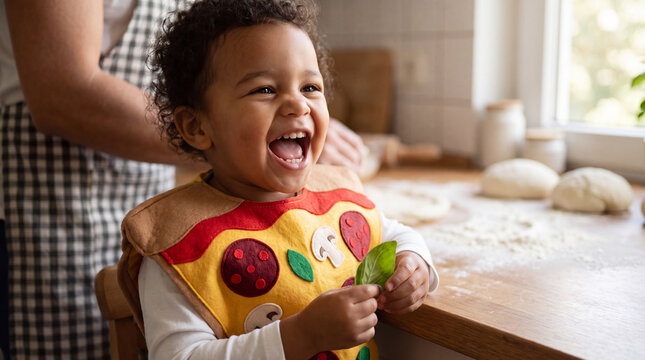 A mixed-race toddler in a pizza costume laughs holding a basil leaf in a home kitchen for National Pizza Month social media posts.