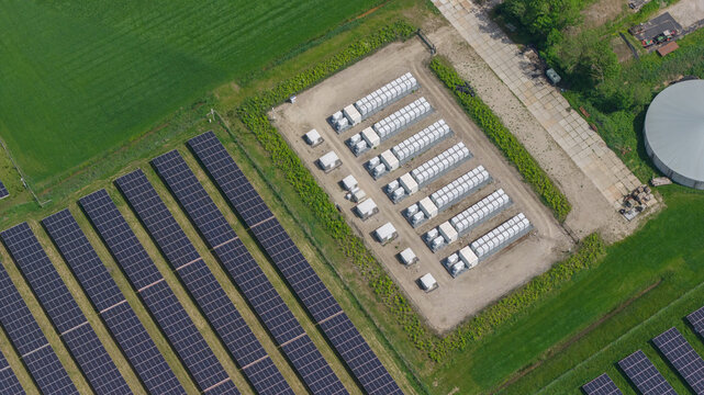 Aerial view of a large solar farm featuring rows of photovoltaic panels and a central battery energy storage system with white containers in Wanneperveen, Overijssel, Netherlands.