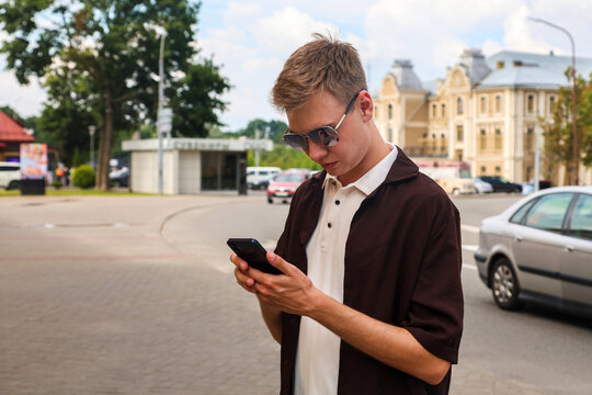 A young blond man in brown clothes stands on the street of a European city with a phone in his hand.