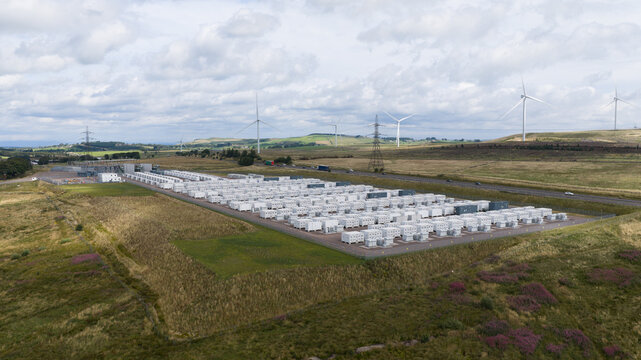 Aerial view of Coalburn BESS facility featuring rows of white battery storage containers set within a grassy landscape with wind turbines in the background in Lanark, Scotland, United Kingdom.