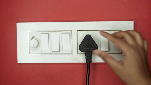 Close up of young adult woman hand holding and plugging black electrical plug in wall outlet socket at home. Electrical equipment wires and power strips on the red wall.