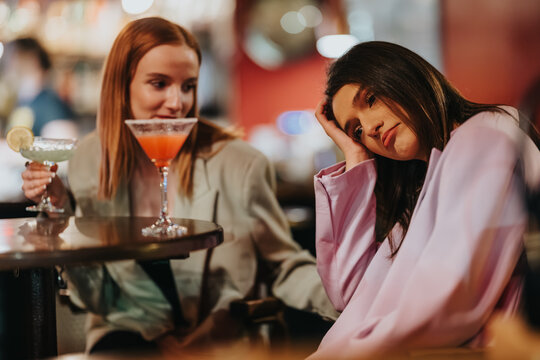 Two girls sit at a bar, one looking sad while the other holds cocktails. A friend offers quiet company during a subdued night out.