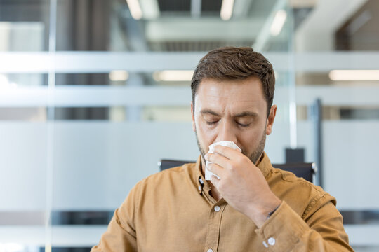 Male employee feeling unwell with a cold or allergy, blowing his nose with a tissue while working in a modern office, symbolizing illness and health concerns in the workplace
