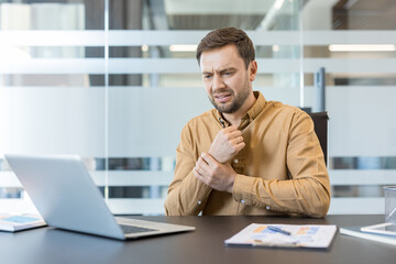 Obraz premium Businessman experiencing acute wrist pain and discomfort while sitting at an office desk and working on a laptop, indicating potential carpal tunnel syndrome or a repetitive strain injury