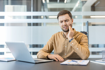 Obraz premium Man experiencing dental pain and discomfort while sitting at his office desk, holding his cheek and trying to focus on his laptop during a busy workday