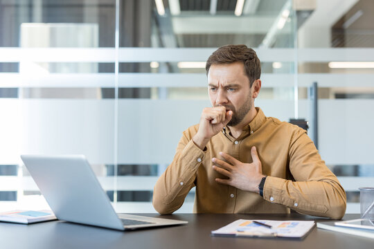 Man feeling unwell and coughing into fist, experiencing illness symptoms while working at his desk in a modern office, highlighting workplace health and the spread of respiratory viruses