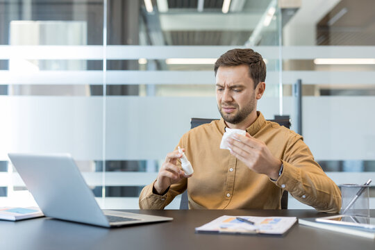 Unwell man experiencing allergy symptoms or a cold, sitting at an office desk with a laptop, holding a nasal spray and a tissue, feeling discomfort while trying to work