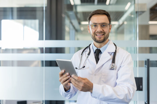 Young male doctor in a white lab coat with stethoscope smiling and holding a digital tablet in a modern clinic, conveying confident, friendly telemedicine and patient care expertise