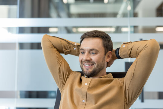 Happy businessman leaning back with hands behind head, smiling in a bright modern office, enjoying a confident, relaxed break after achieving work goals and feeling satisfied and motivated